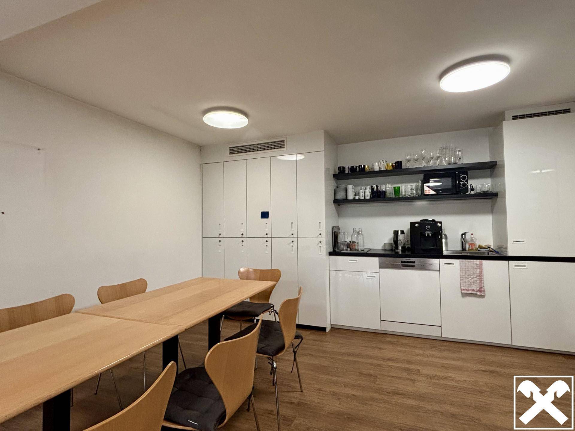 Moderne Teeküche mit langer Holztafel, weißen Schränken und integrierten Geräten wie Geschirrspüler und Mikrowelle.