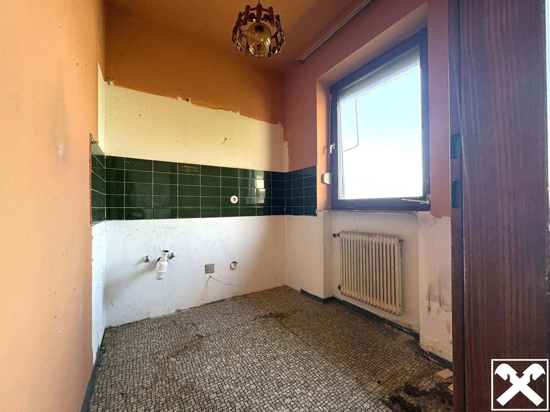 Leerer Raum mit Fenster, Heizkörper und alten Fliesen, der Renovierungsbedarf aufweist.