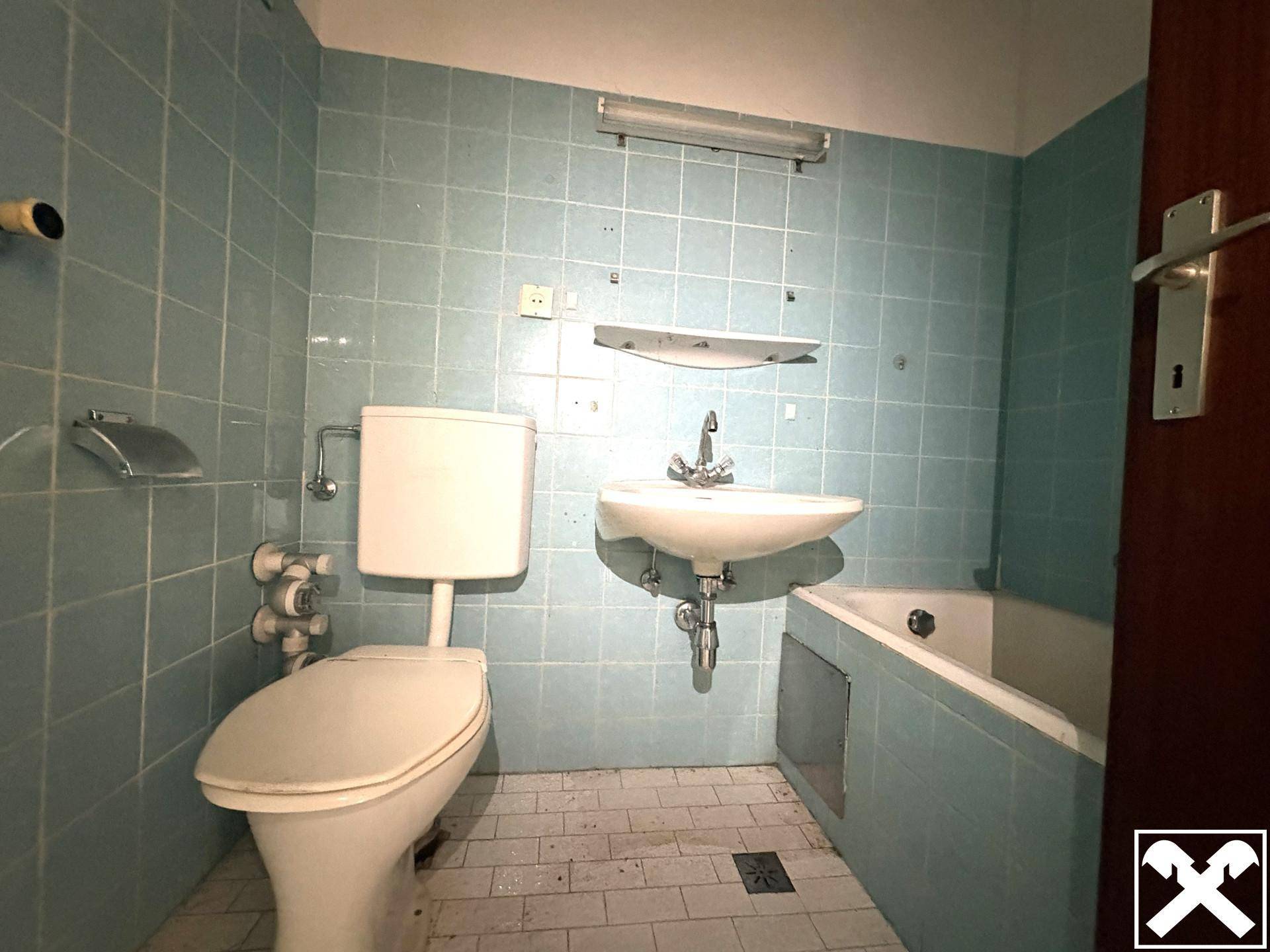 Badezimmer mit hellblauen Fliesen, Toilette, Waschbecken und einer Badewanne, in originalem Zustand.