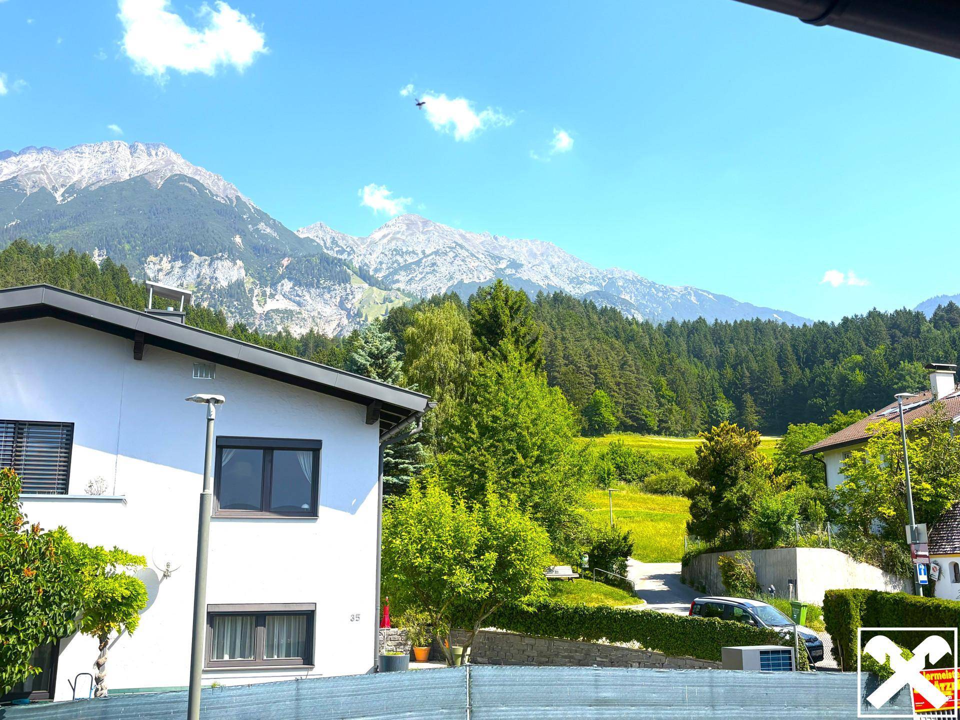 Hausfassade mit Blick auf grüne Bäume und majestätische Berge unter blauem Himmel.