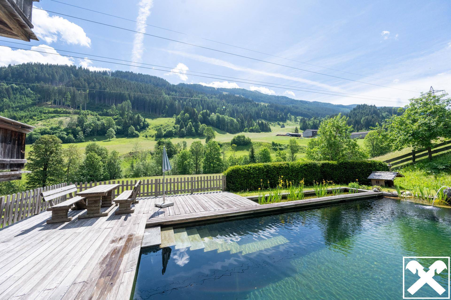 Naturpool mit Holzdeck und Sitzbereich, umgeben von grünen Hügeln und alpiner Landschaft.
