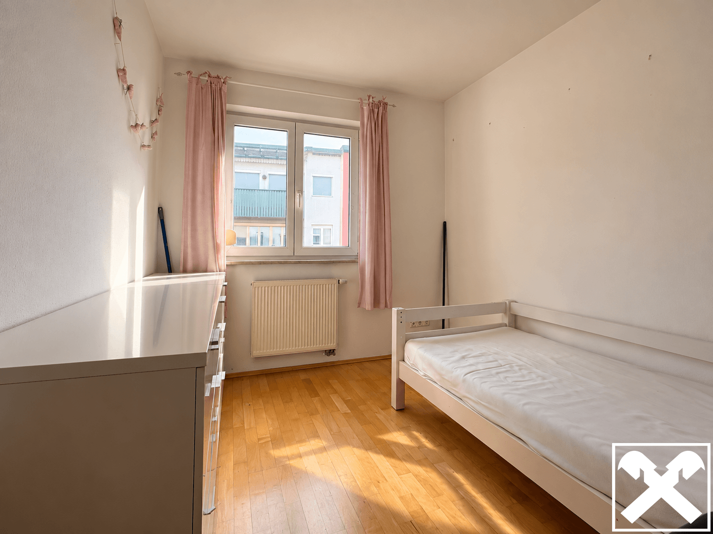 Helles Kinder- oder Gästezimmer mit Holzboden, Fenster und einfacher Einrichtung.