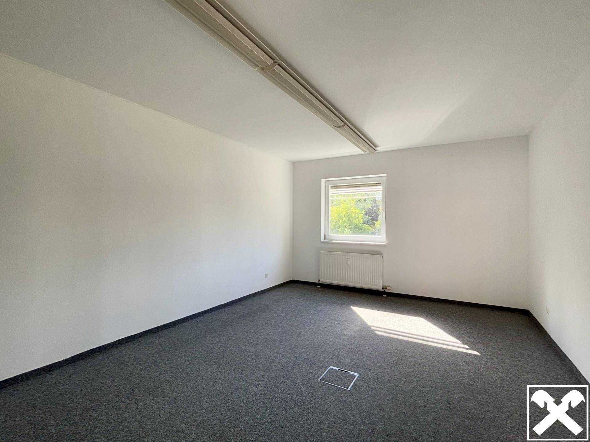 Leeres Zimmer mit grauem Teppichboden, weiß gestrichenen Wänden und einem Fenster mit Heizkörper.