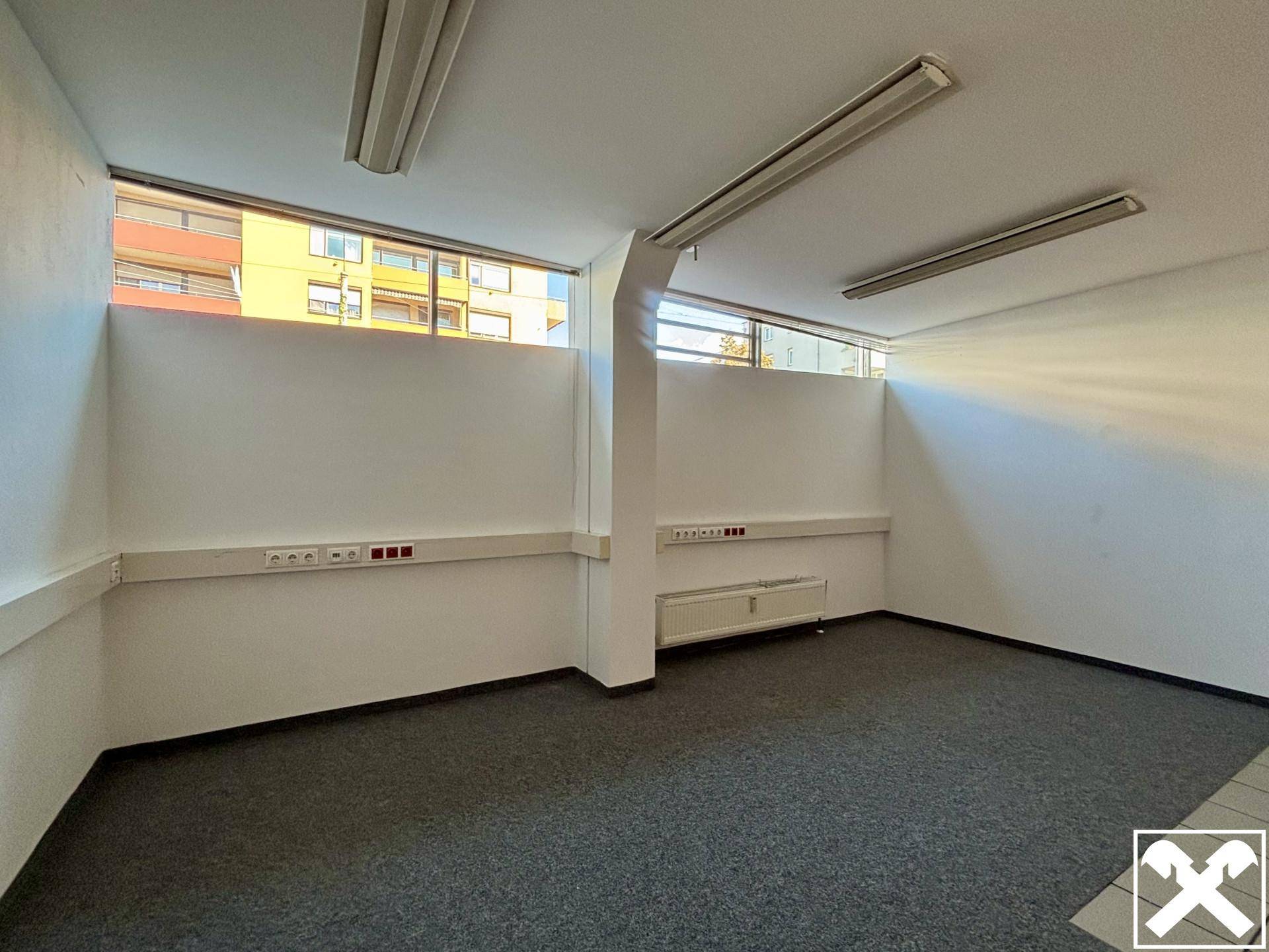Funktionaler Büroraum mit grauem Teppichboden, weißen Wänden und hochgesetzten Fenstern.