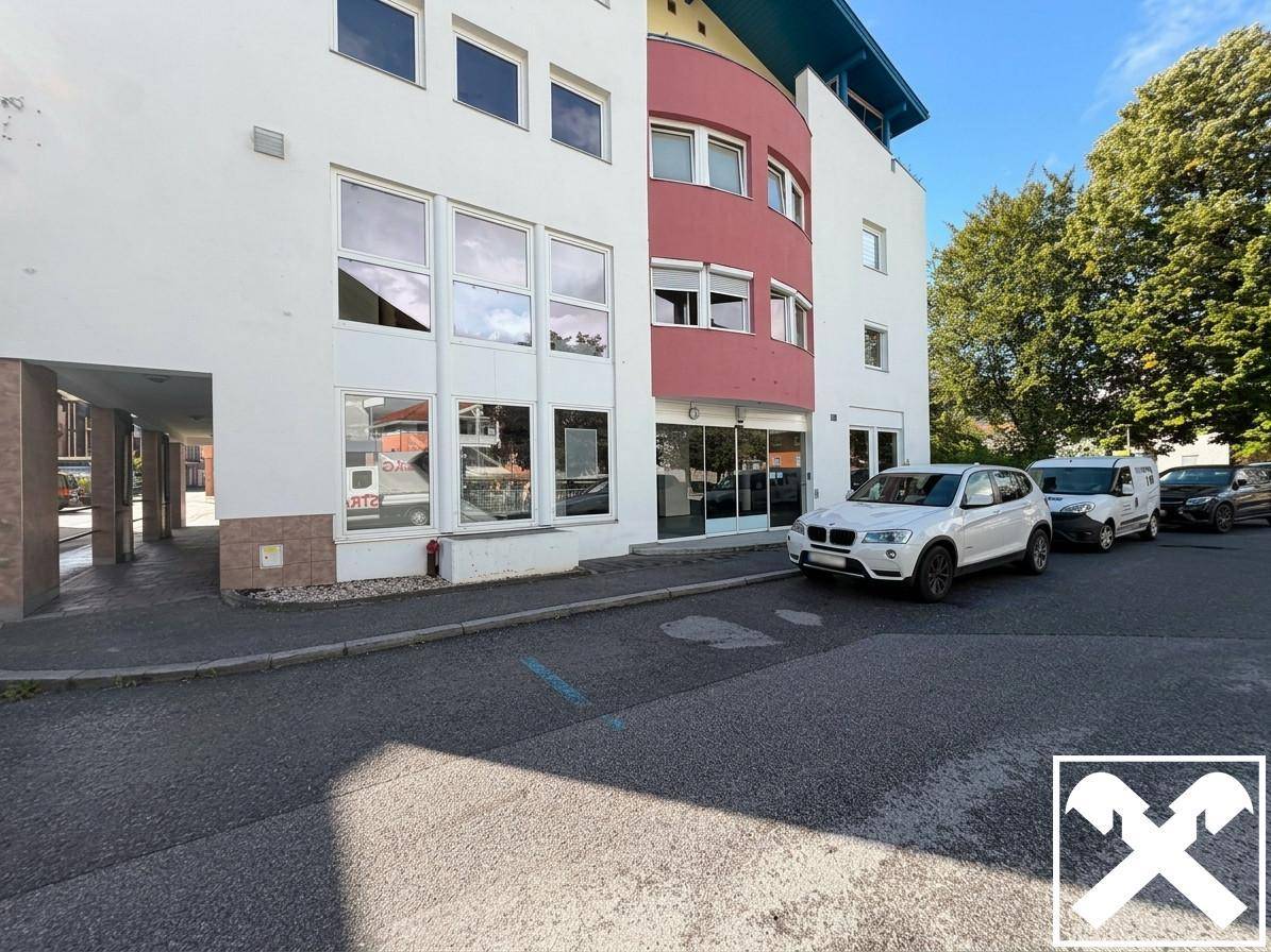 Moderne Gebäudefassade mit großen Fensterfronten, roten Akzenten und Parkplätzen davor.