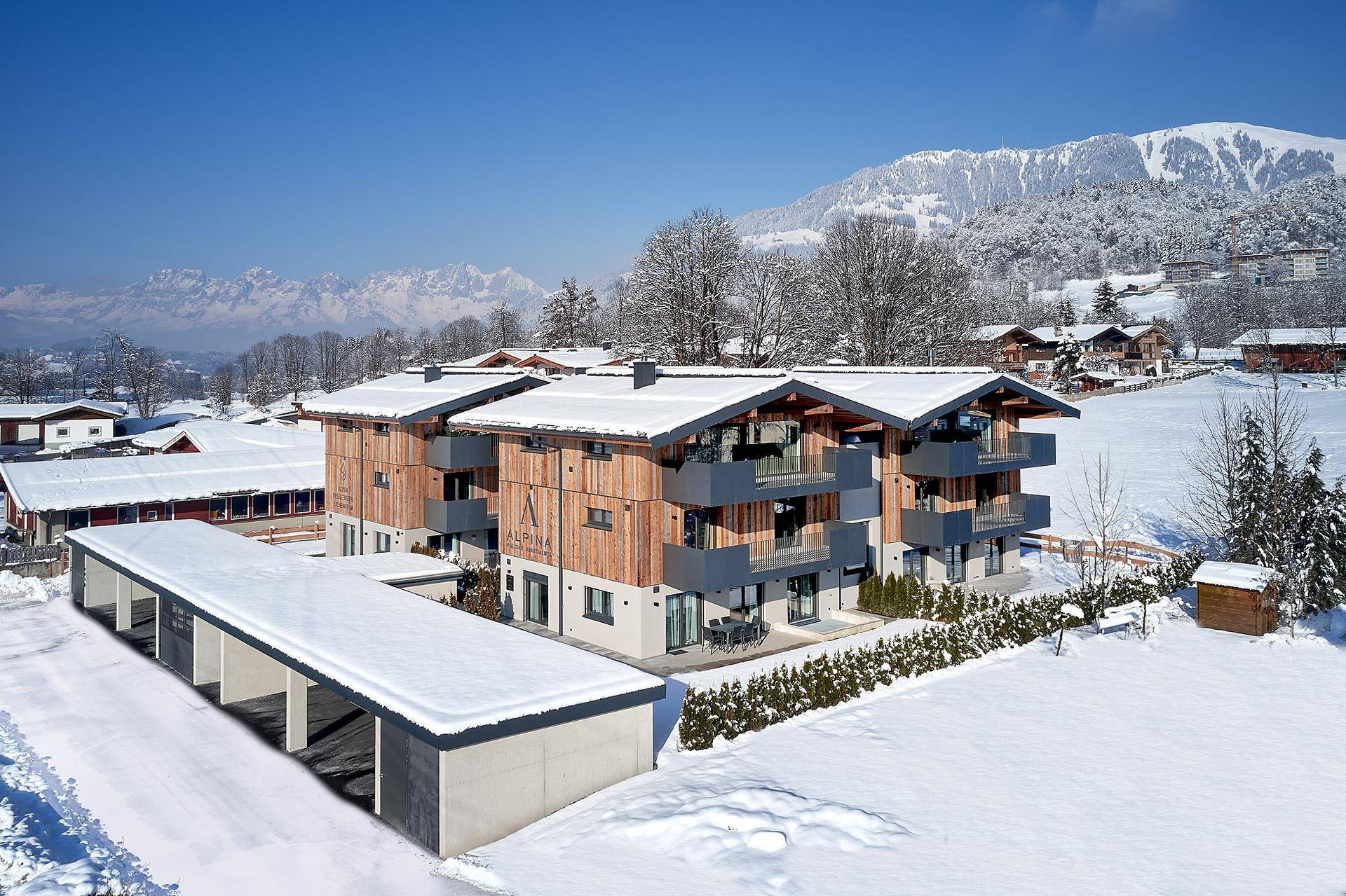 Neues Apartmenthaus mit Carport in verschneiter Alpenlandschaft und Bergblick an einem sonnigen Tag.