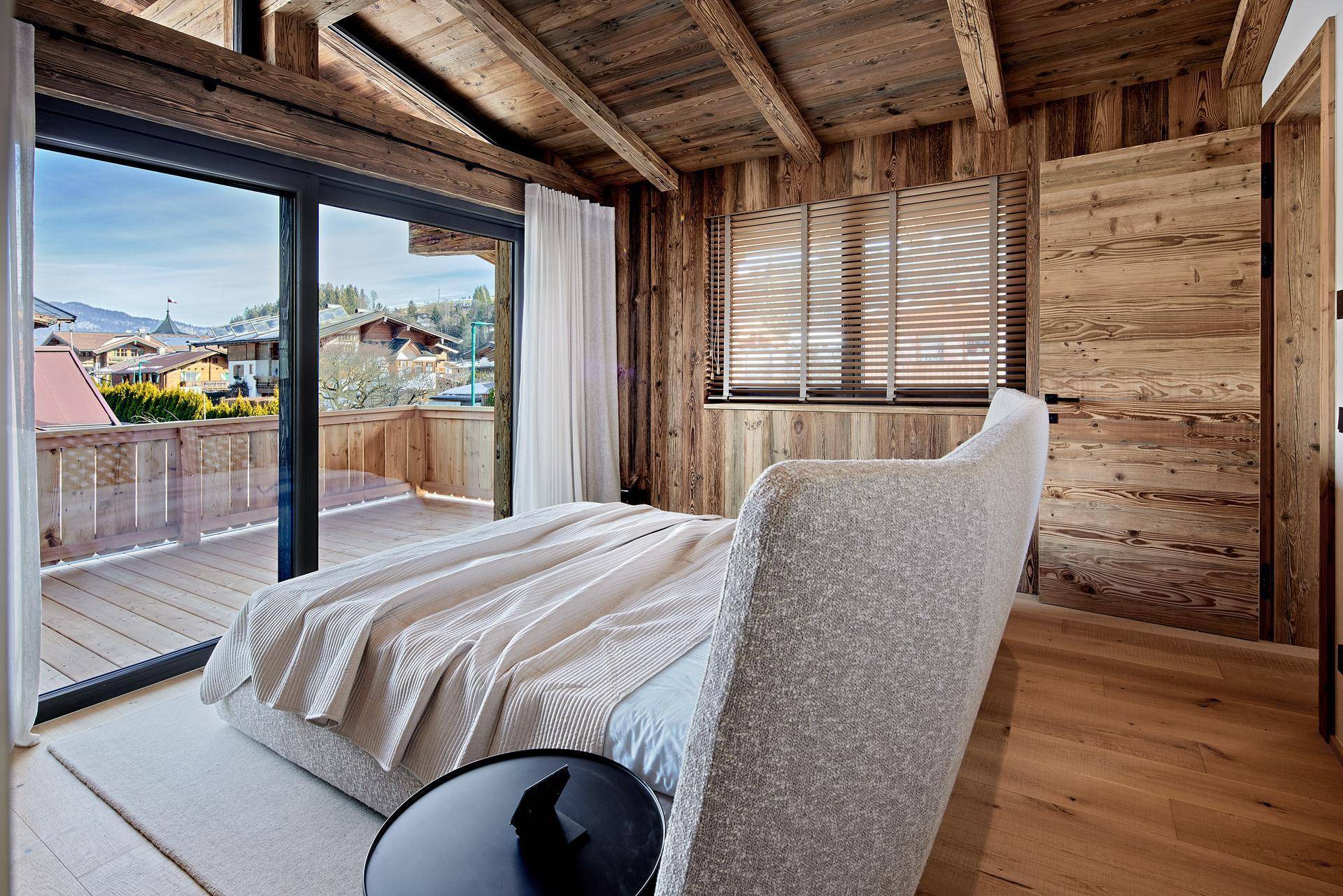 Helles Schlafzimmer mit Holzbalkendecke, großen Fenstern und Zugang zu einem sonnigen Balkon.