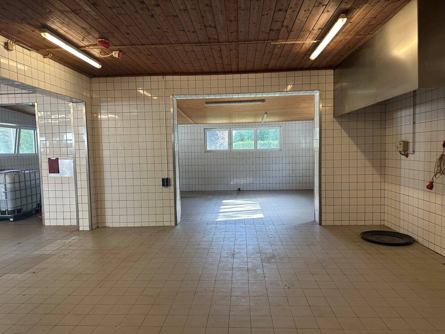 Gewerbliche Küche mit weißen Fliesenwänden, Fliesenboden und einer Holzdecke, gut beleuchtet durch Fenster.