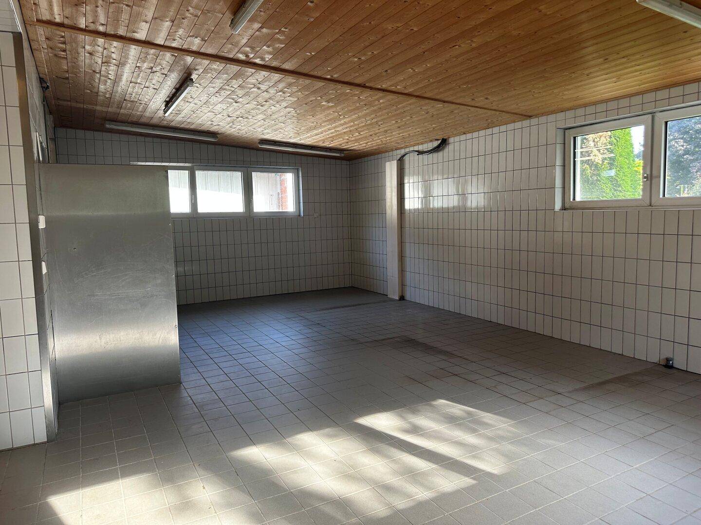 Geräumige Gewerbeküche mit weißen Fliesenwänden, Fliesenboden und einer warmen Holzdecke, hell durch Fenster.