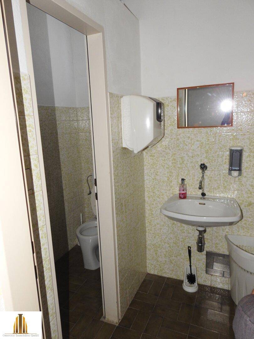 Einfache Toilette mit Waschbecken und hellgrünen Fliesen, angrenzend an einen weiteren WC-Bereich.