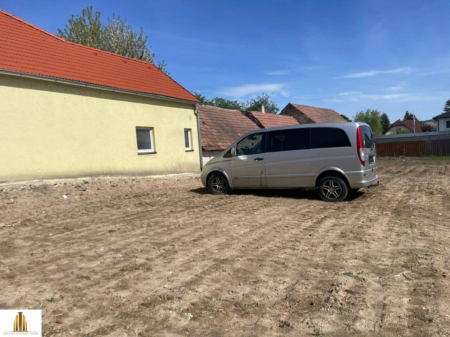 Freies Grundstück mit geparktem Fahrzeug und angrenzendem Gebäude mit Ziegeldach unter blauem Himmel.