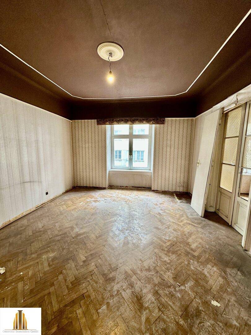 Geräumiges Zimmer mit abgenutztem Fischgrätparkett und hohen Decken, bereit für eine umfassende Renovierung.