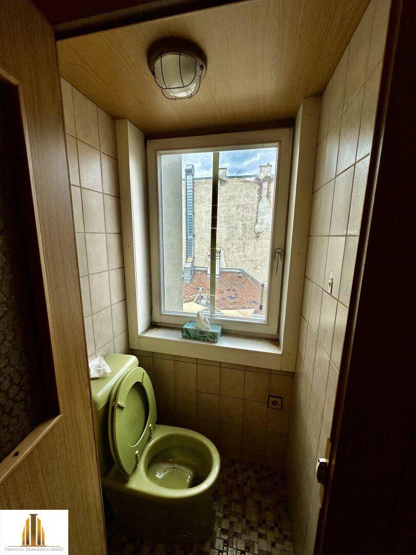 Kleines WC mit grünem WC und Fenster, gefliest in beigen Tönen, in originalem Zustand.