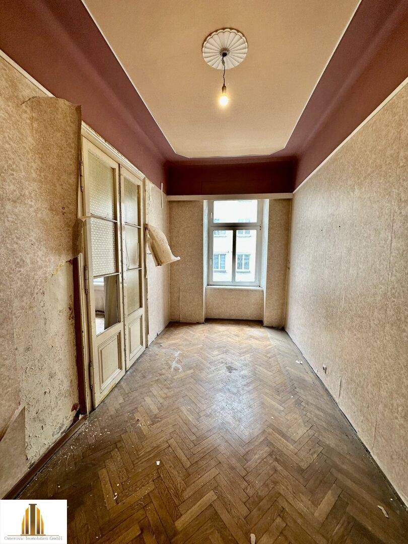 Leeres Zimmer mit abgenutztem Fischgrätparkett und hohen Decken, ideal zur Sanierung und Neugestaltung.