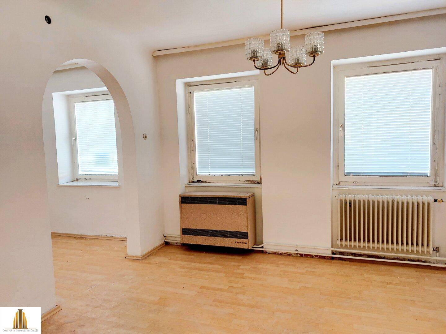 Helles Wohnzimmer mit Parkettboden, zwei Fenstern und einem dekorativen Bogen zu einem weiteren Raum.