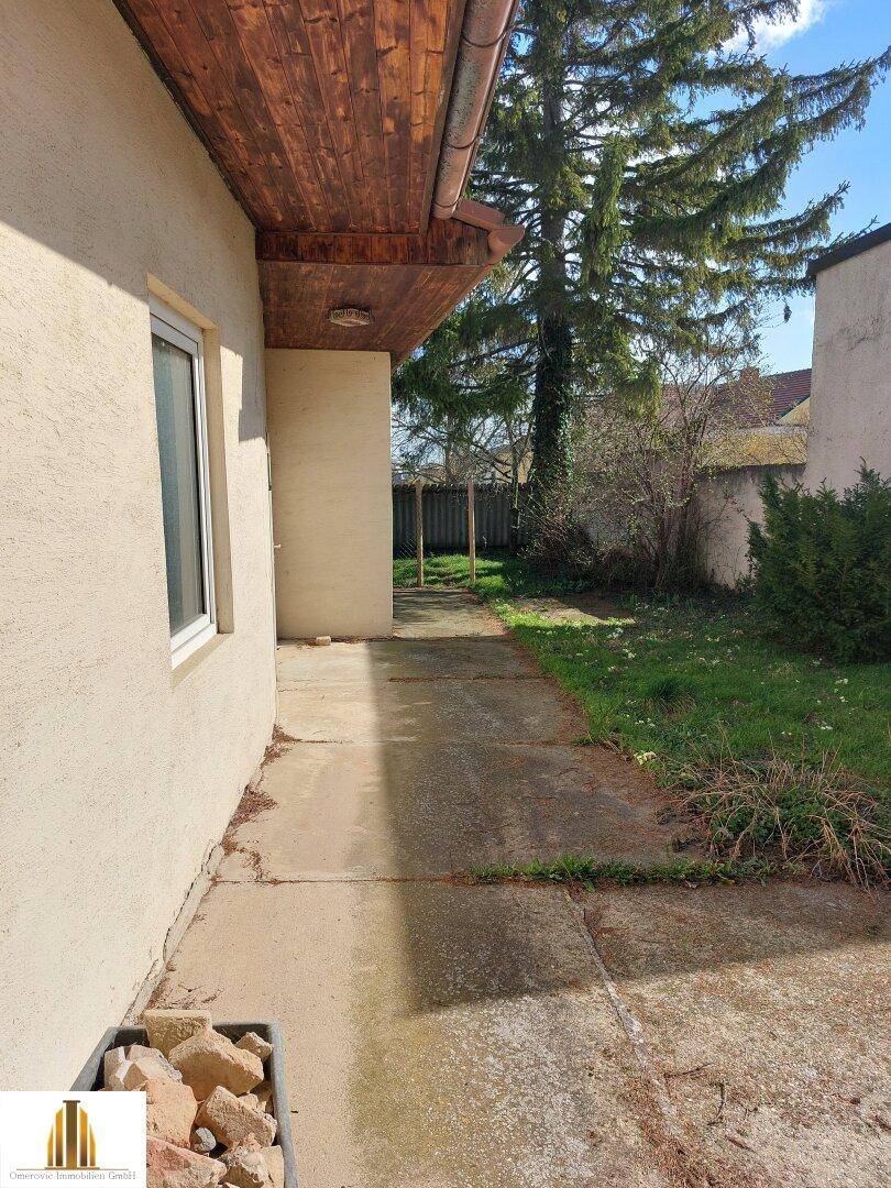 Seitenansicht des Hauses mit gepflastertem Weg und angrenzender Rasenfläche unter einem großen Baum.