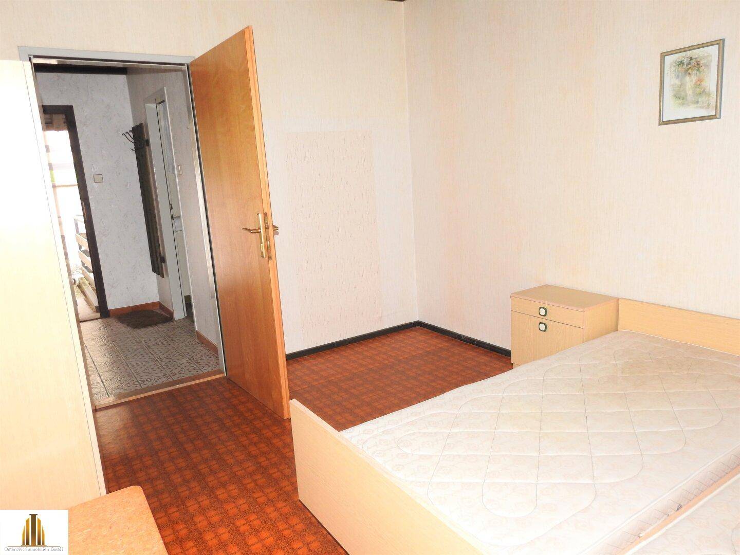 Schlafzimmer mit Bett, Nachttisch und Blick in den Flur, einfach eingerichtet.