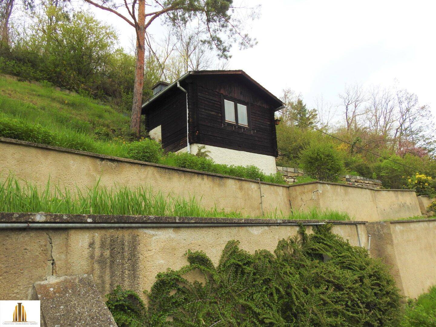 Kleines Holzhaus am Hang mit grüner Umgebung und Stützmauern, idyllisch gelegen.