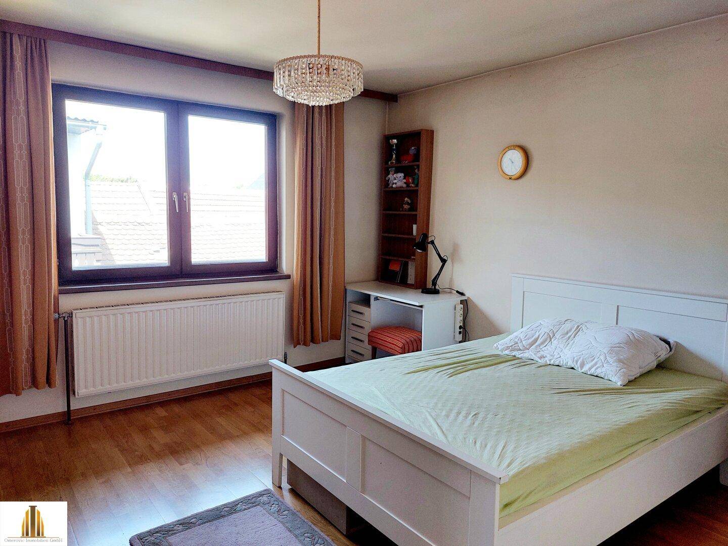 Schlafzimmer mit Holzboden, Schreibtisch und Fenster mit Vorhängen.