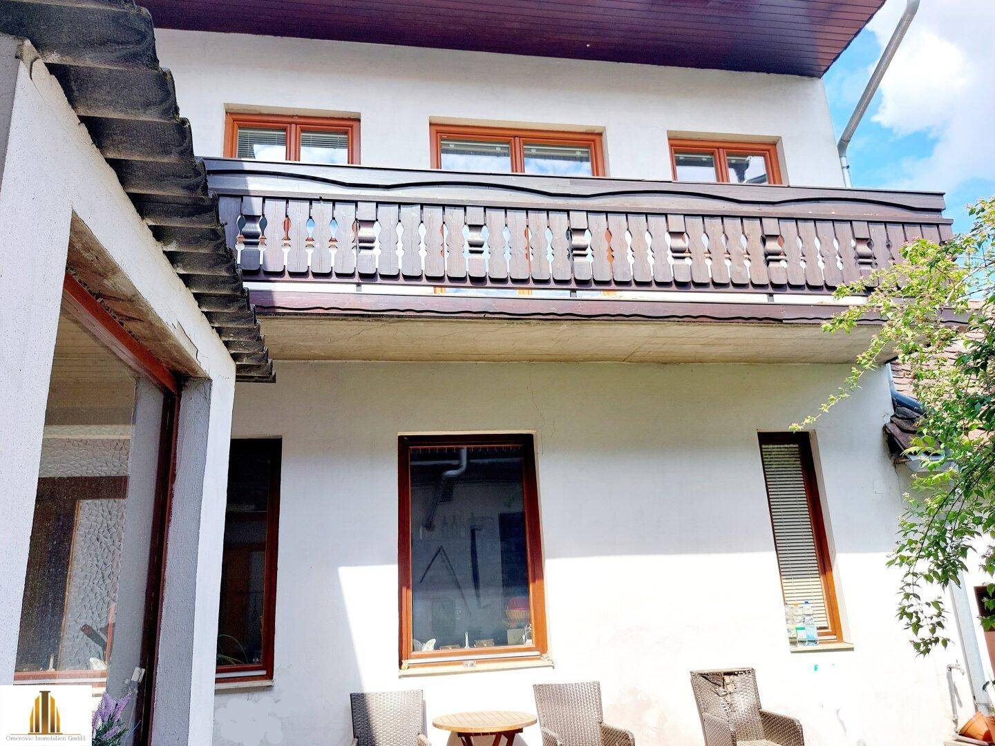 Ansicht der Hausfassade mit einem Balkon im Obergeschoss und einem Sitzbereich im Erdgeschoss.