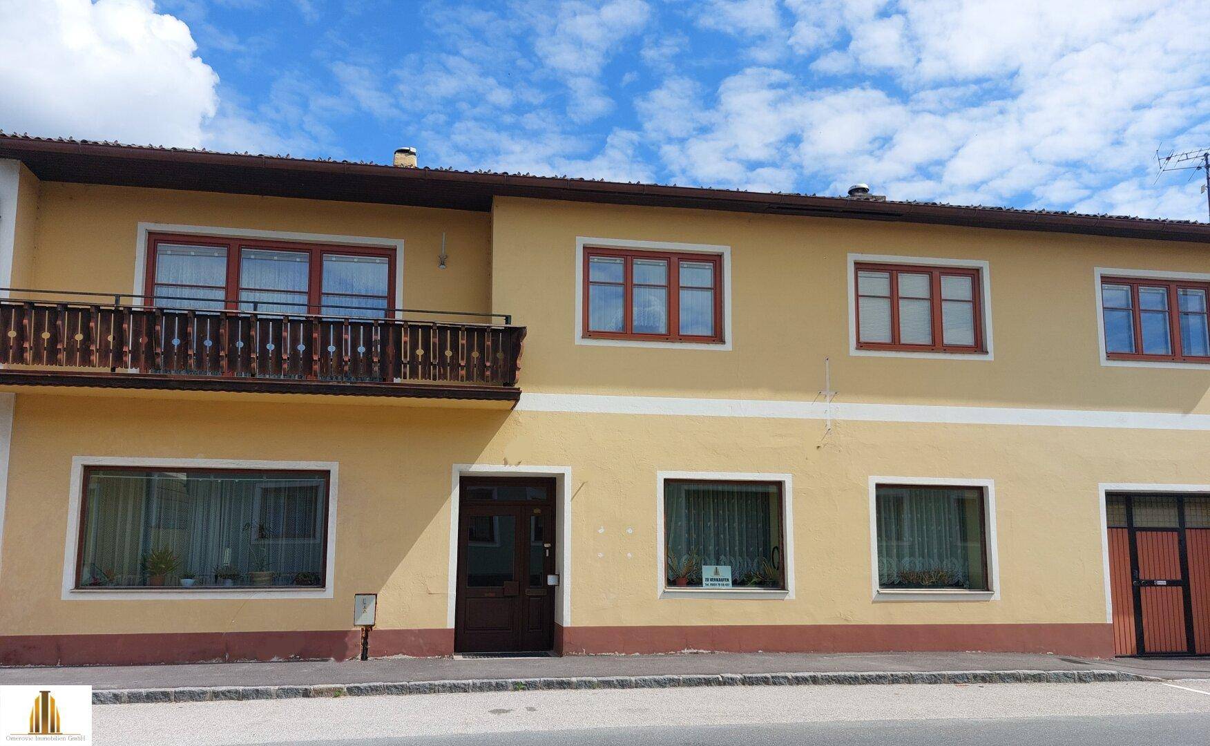 Gelbe Fassade eines Mehrfamilienhauses mit Holzbalkon und braunen Fensterrahmen unter blauem Himmel.