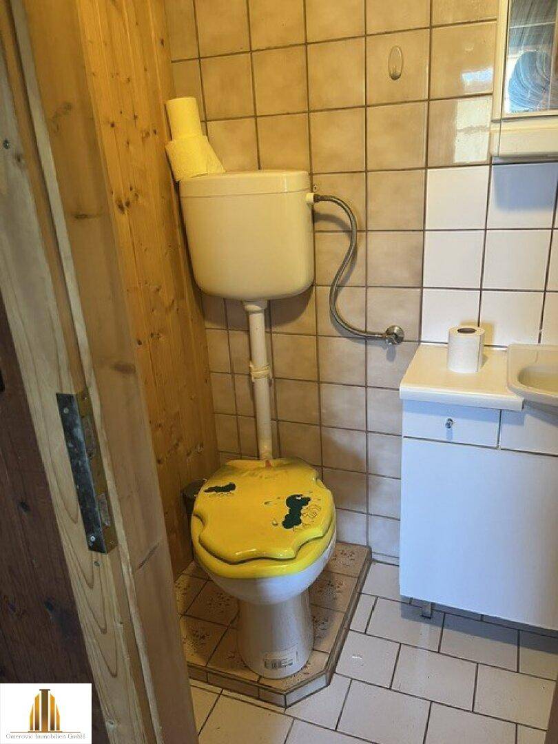 Separate Toilette mit gelbem Toilettensitz und weißen Wandfliesen.