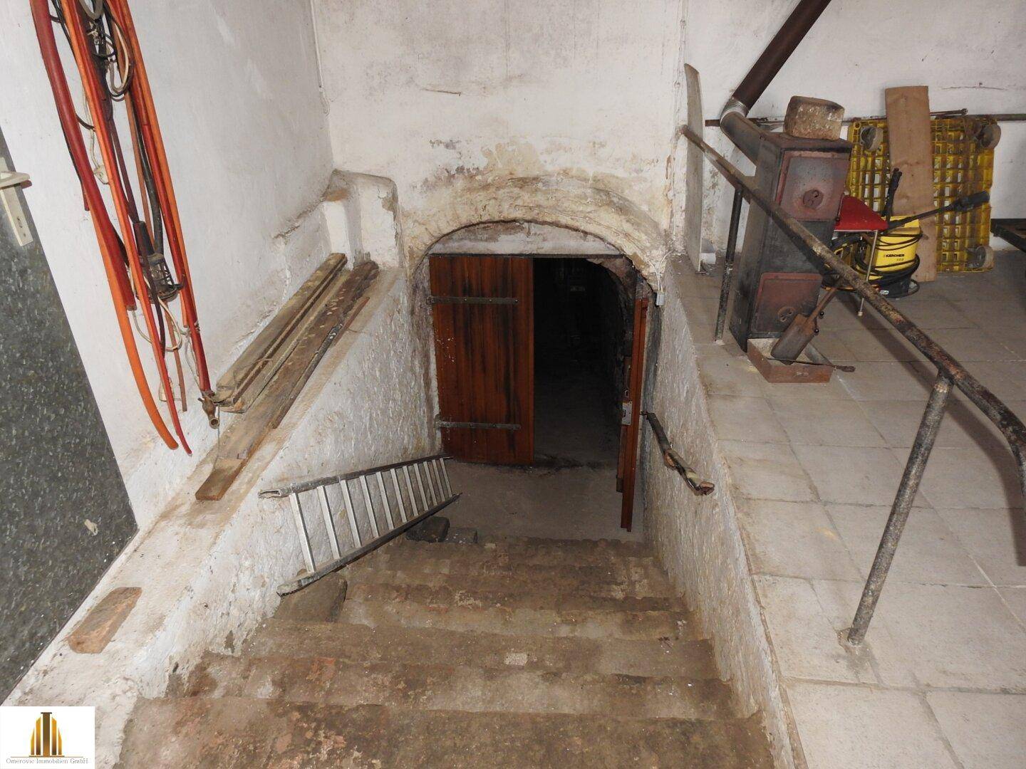 Steile Steintreppe führt hinab in einen dunklen, rustikalen Kellerraum mit Holztür.