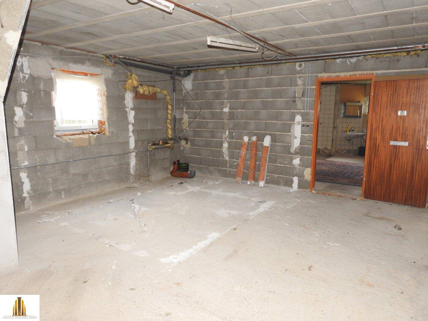 Unausgebauter Kellerraum mit Betonwänden, kleinem Fenster und Zugang zu einem weiteren Raum.