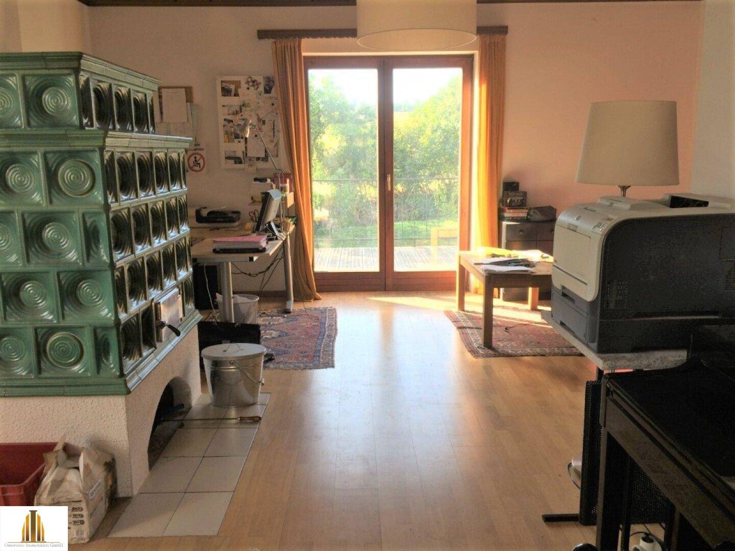 Helles Wohnzimmer mit Kachelofen, Holzboden und Zugang zur Terrasse mit Gartenblick.