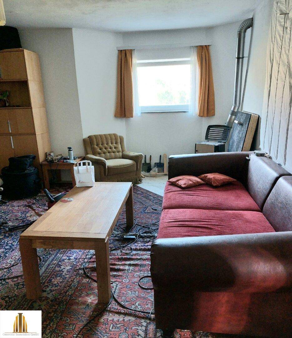 Gemütliches Wohnzimmer mit Teppichboden, Sofa, Sessel und einem Fenster mit Vorhängen.