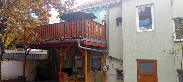 Hinteransicht des Gebäudes mit einem Holzbalkon und einer überdachten Terrasse im Erdgeschoss.