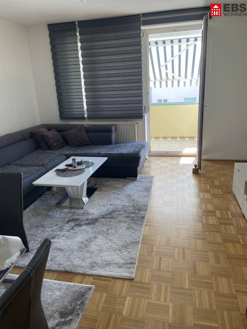 Gemütliches Wohnzimmer mit grauem Ecksofa, Parkettboden und Zugang zu einem Balkon.