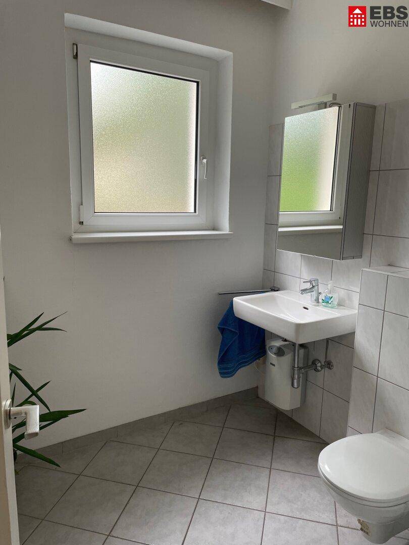 Helles Badezimmer mit Fenster, modernem Waschbecken, wandhängender Toilette und grauen Fliesen.
