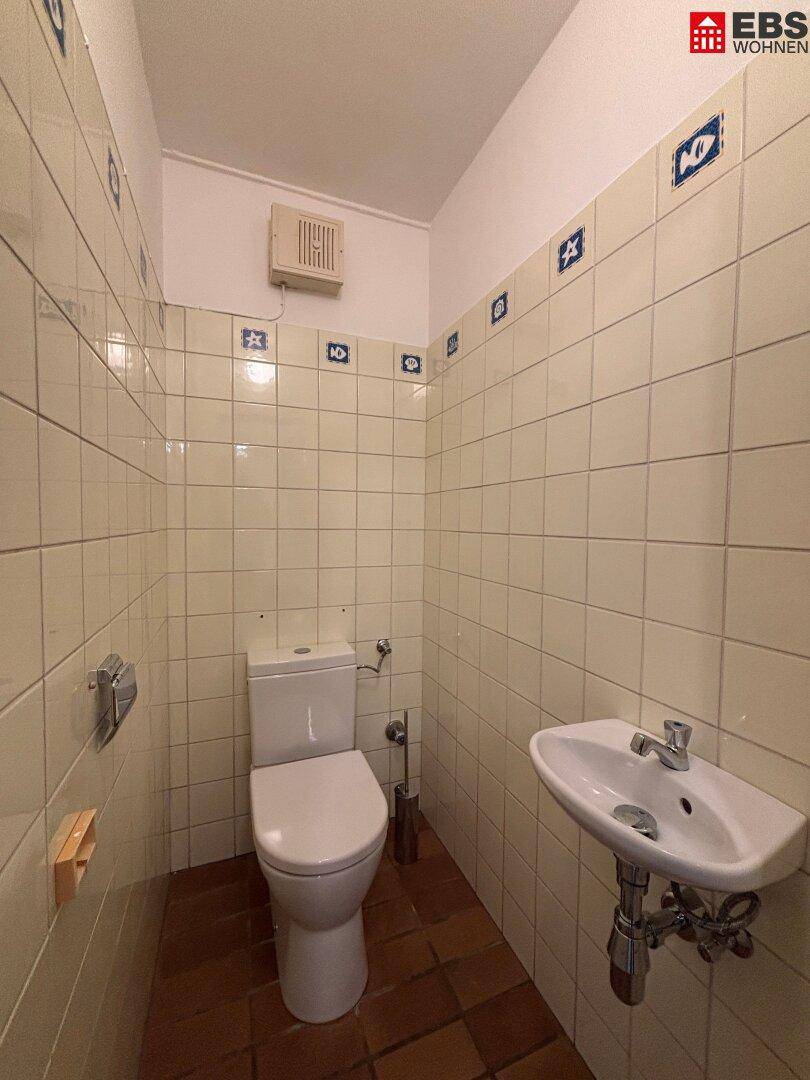Einfaches Gäste-WC mit hellen Fliesen, Toilette und kleinem Waschbecken.
