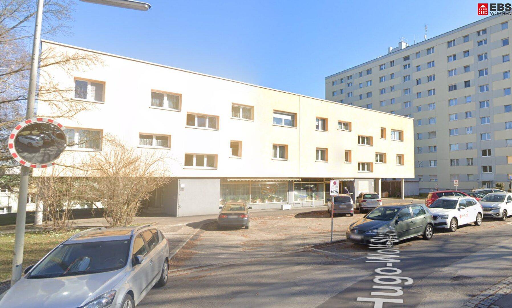 Weitläufige Straßenansicht des Gebäudes mit parkenden Autos und umliegender Bebauung.