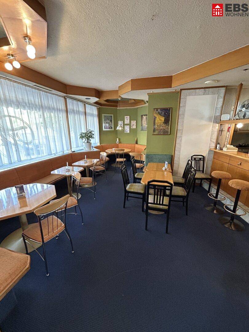 Geräumiger Café-Innenraum mit zahlreichen Tischen, Stühlen und einer langen Sitzbank am Fenster.