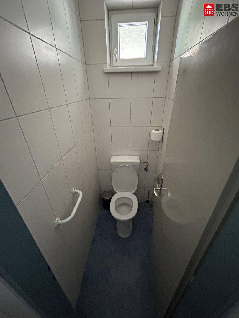 Toilette mit Haltegriffen an der Wand, WC und einem Fenster für Belüftung.