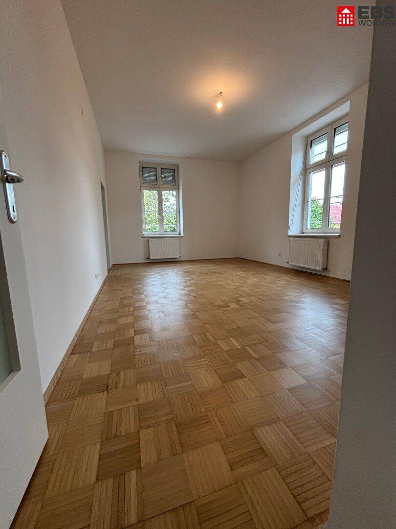 Geräumiges, leeres Wohnzimmer mit klassischem Parkettboden und zwei großen Fenstern.