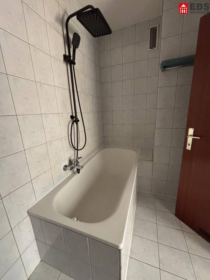 Badezimmer mit Badewanne, Duscharmatur und weißen Fliesen an Wänden und Boden.