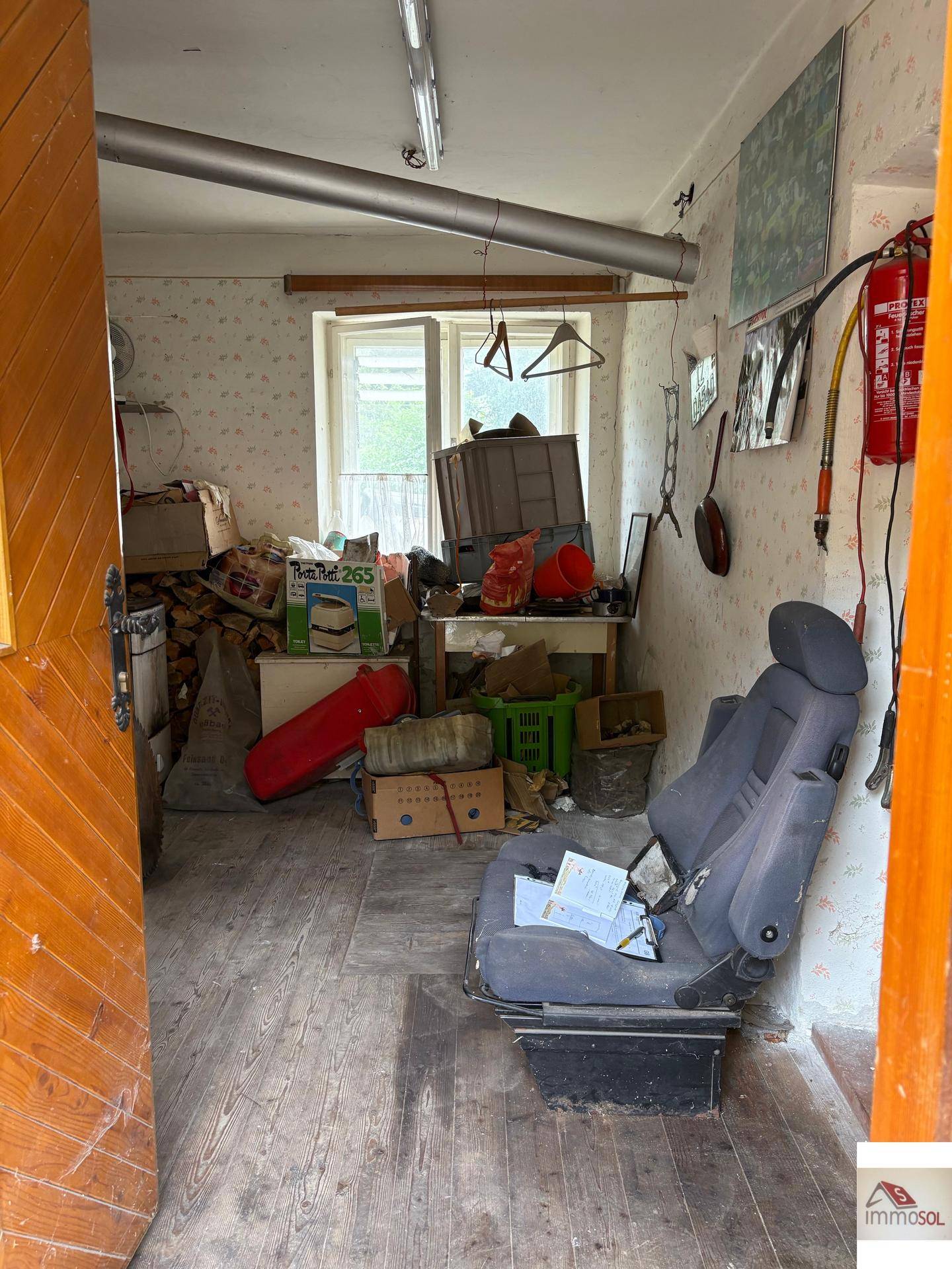 Überfüllter Abstellraum mit altem Mobiliar, Tapeten und einem Fenster, das Tageslicht hereinlässt.