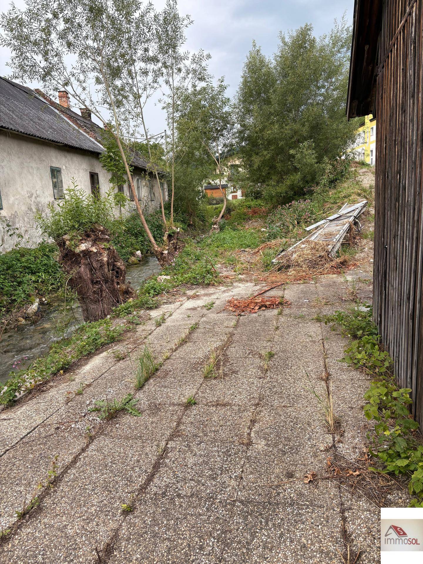Gepflasterter Weg neben einem alten Gebäude und einem Bachlauf, umgeben von Bäumen und Sträuchern.