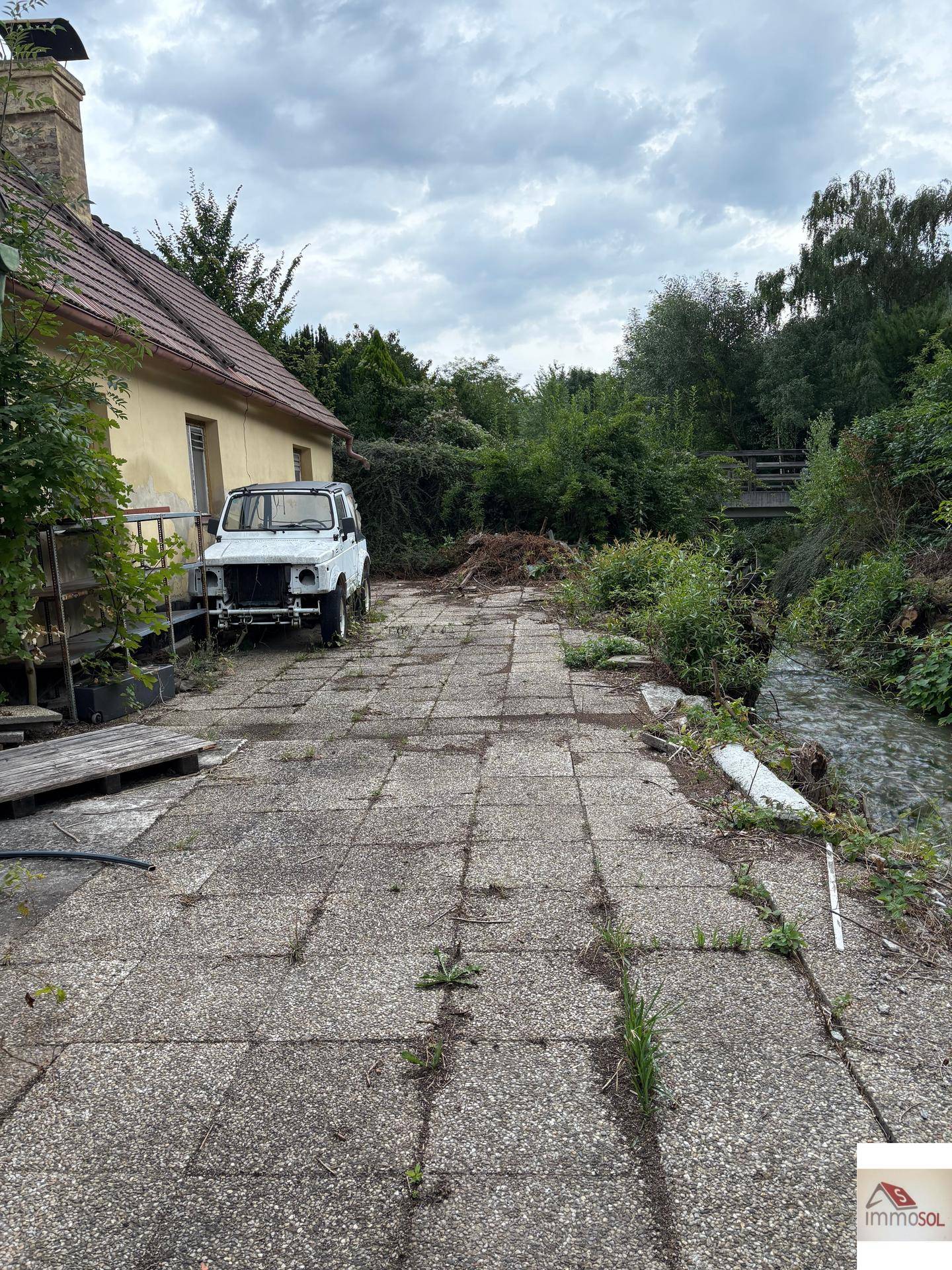 Gepflasterter Hofbereich neben einem Haus mit einem alten Auto und viel Grünbewuchs.