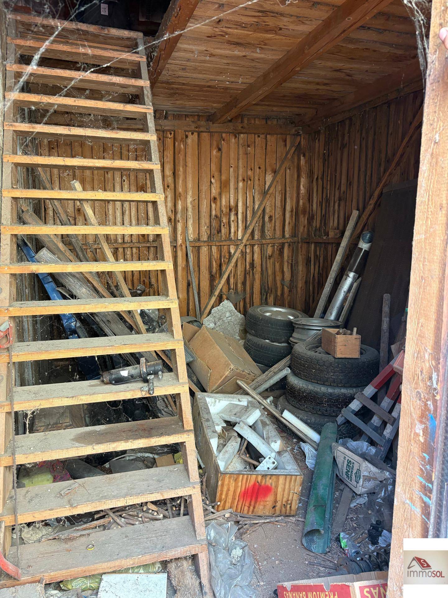 Innenansicht eines Holzschuppens mit einer Leiter und verschiedenen gelagerten Gegenständen und Reifen.