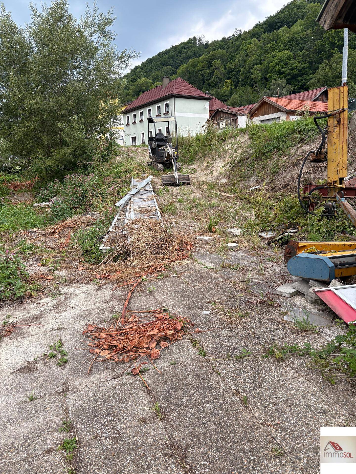 Geräumtes, leicht abschüssiges Grundstück mit gepflastertem Weg und einem Gebäude im Hintergrund.