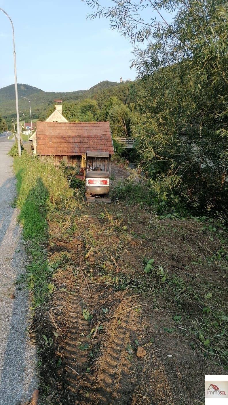 Freigelegtes Grundstück neben einer Straße mit einem kleinen Gebäude und bewaldeten Hügeln im Hintergrund.