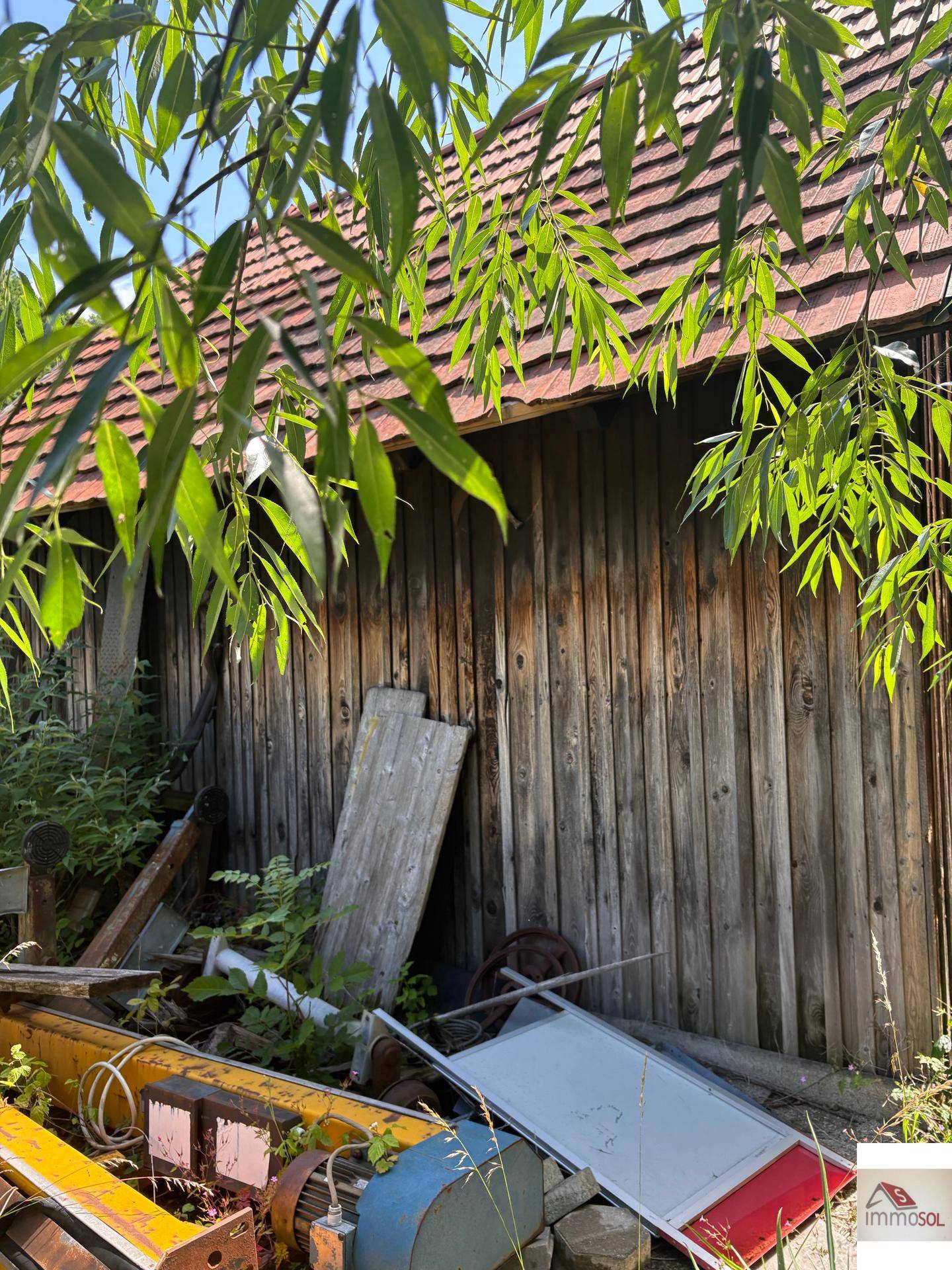 Rustikale Holzscheune mit verwitterten Brettern, umgeben von üppiger Vegetation und alten Materialien.