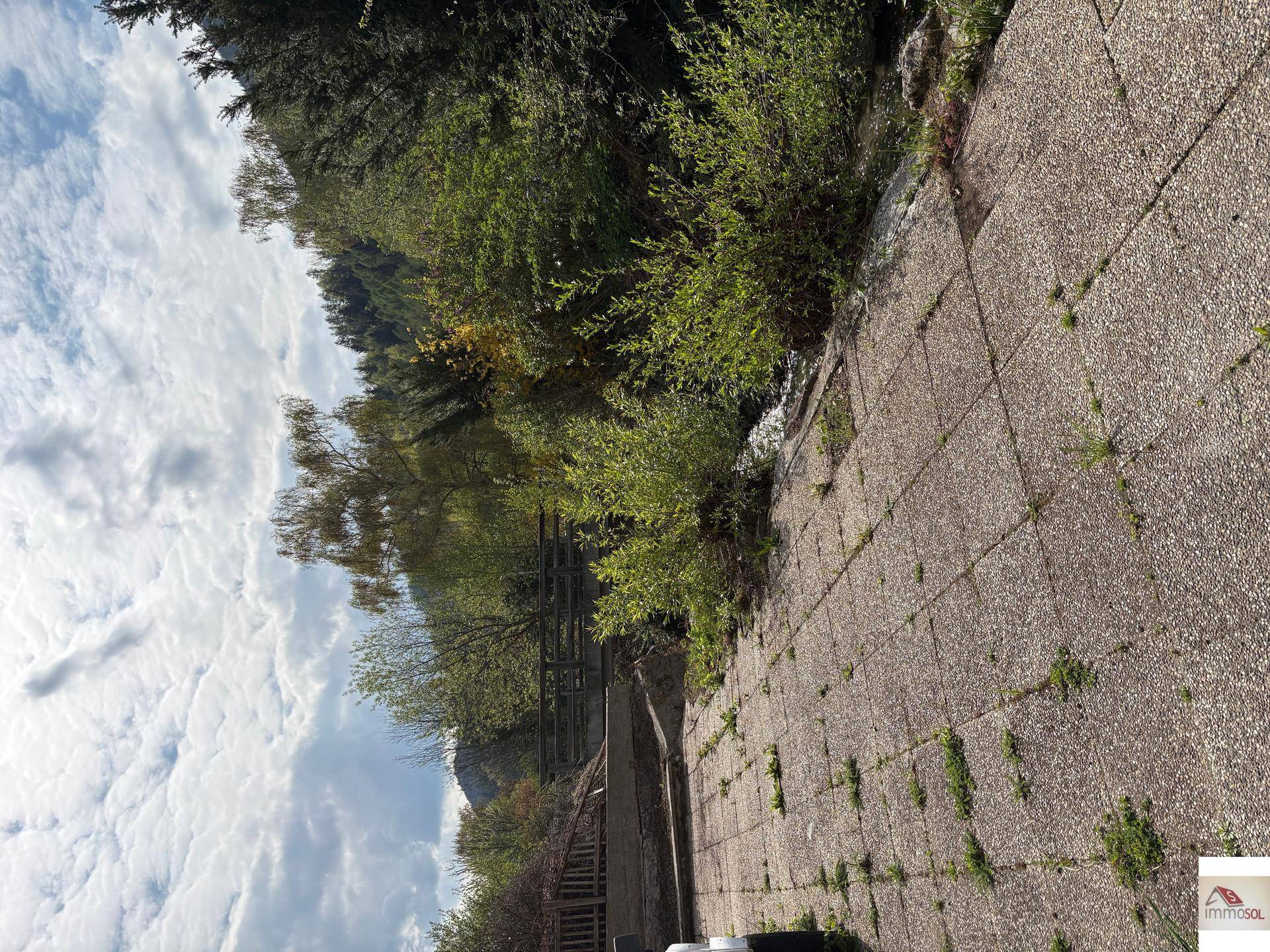 Gepflasterter Bereich am Bachlauf mit üppiger Vegetation und bewölktem Himmel.