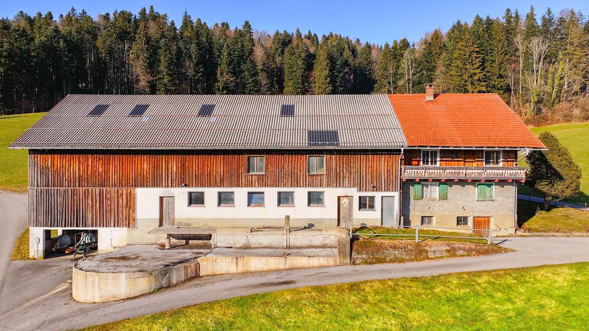 Seitenansicht eines Bauernhauses mit Holzfassade, Ziegeldach und Solarmodulen in ländlicher Umgebung.
