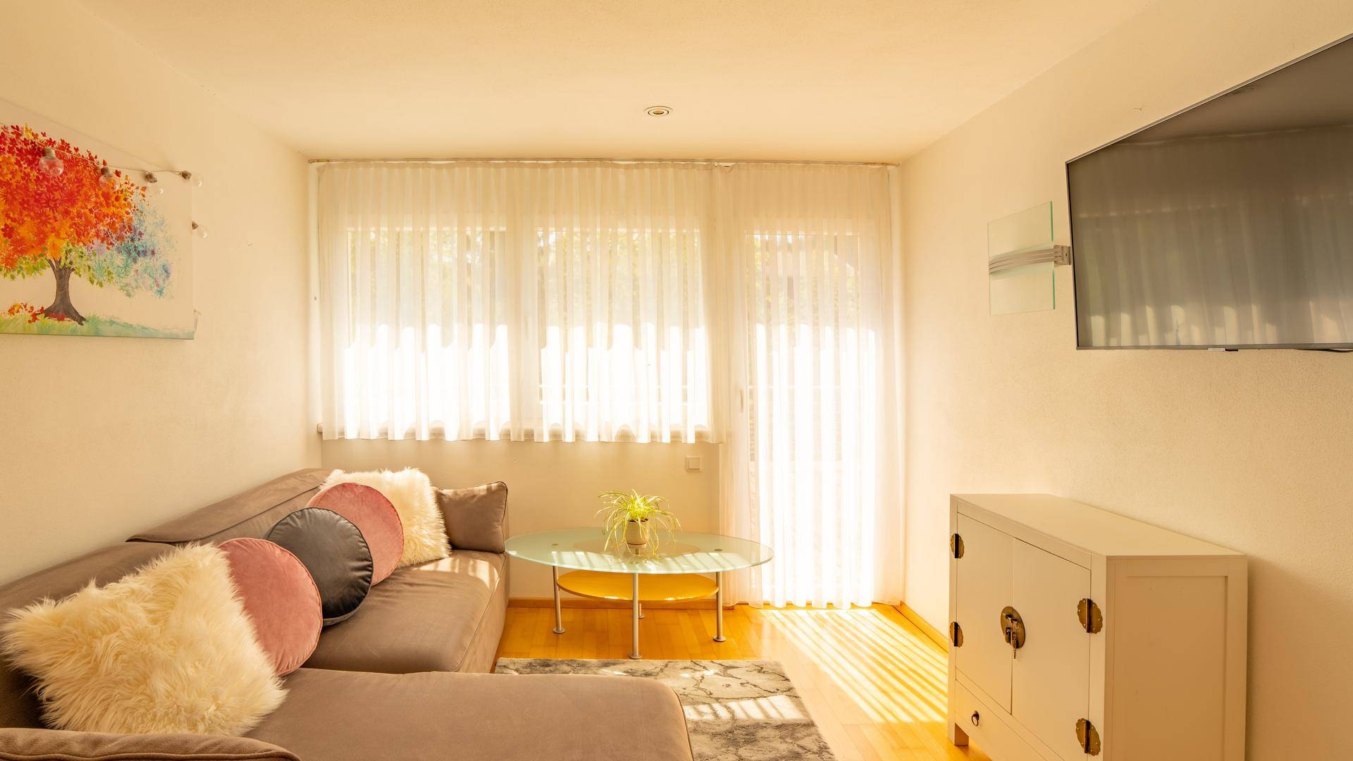 Helles Wohnzimmer mit beigem Sofa, Fernseher und großen Fenstern mit transparenten Vorhängen.