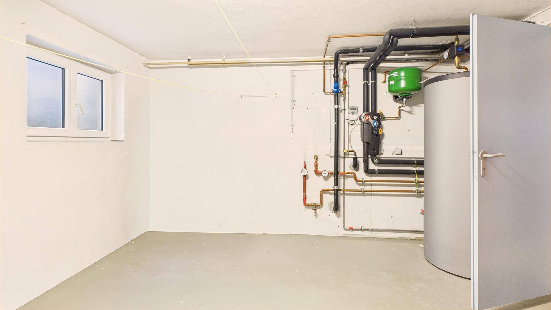Technikraum im Keller mit Heizungsanlage, Rohren und einem Fenster für natürliche Belichtung.